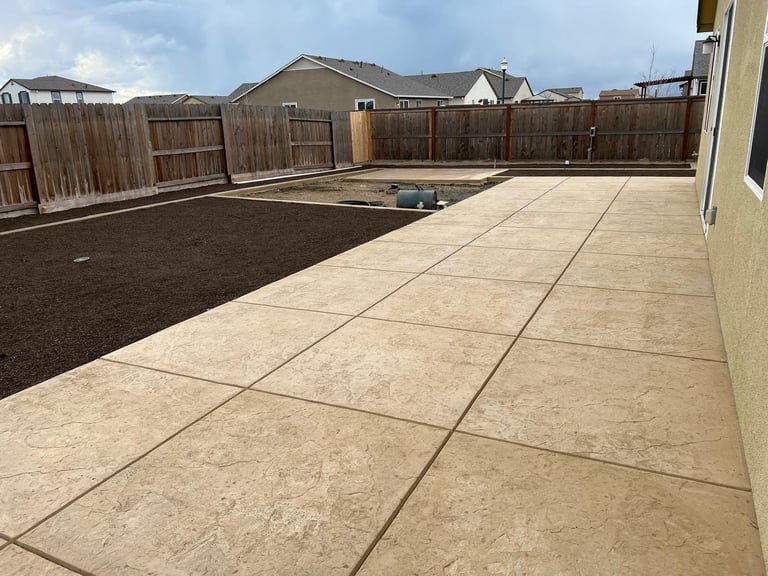 Spacious backyard with concrete patio on right side, empty dirt yard on left, wooden fence, and residential houses in background
