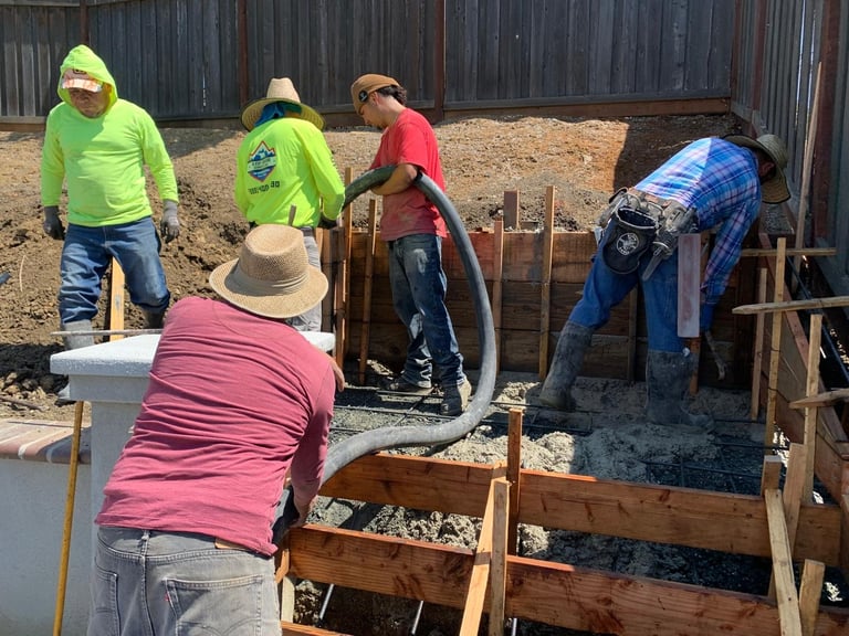 Construction workers in safety vests pouring concrete into a wooden foundation frame at an outdoor construction site