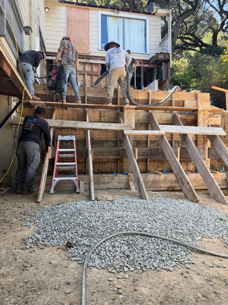 Construction workers building a wooden deck addition to a residential house with metal ladders and gravel visible on the ground
